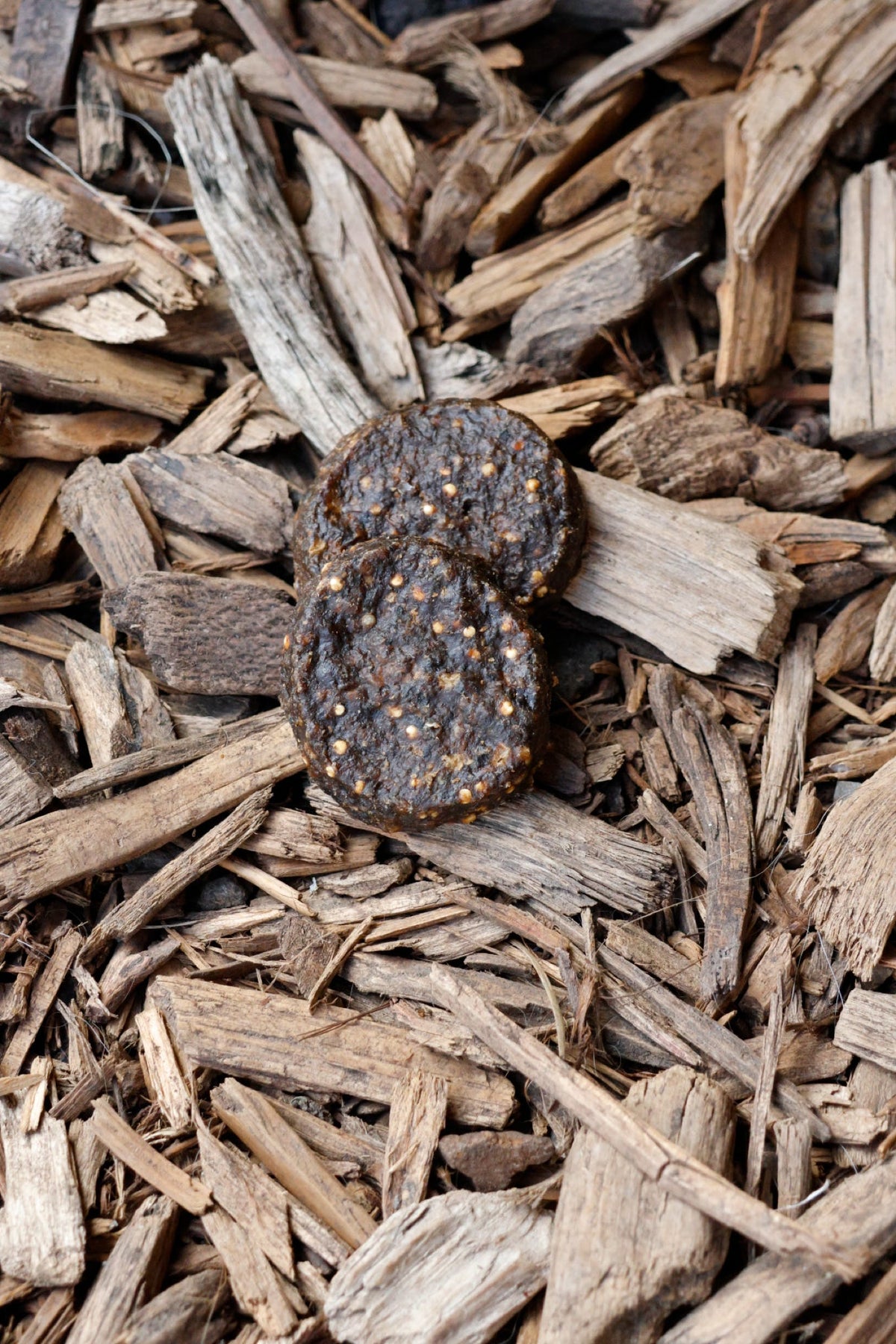 Two dark brown, textured beef burger dog treats stacked on top of natural wood chips.