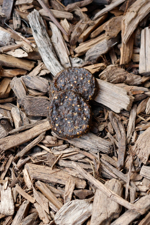 Two dark brown, textured beef burger dog treats stacked on top of natural wood chips.