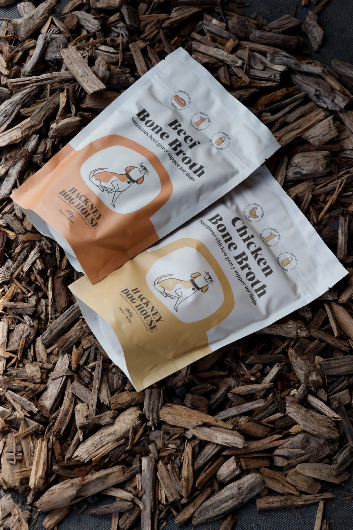 Hackney Dog House Bone Broth pouches, showing Beef Bone Broth and Chicken Bone Broth, displayed on a bed of wood chips.