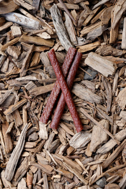 Three red-brown meat sticks crossed in an X shape, with beef, horse and venison varieties, lying on a bed of wood chips.