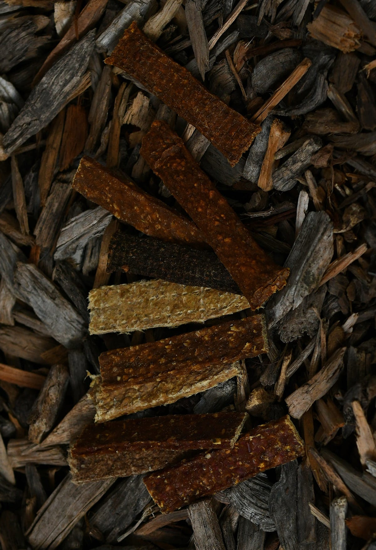 Assorted dried meat strips in various flavours, shown in different colours and textures on a bed of natural wood chips.