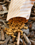 Peanut butter mini bone dog treats spilling from a striped paper bag onto wood mulch.