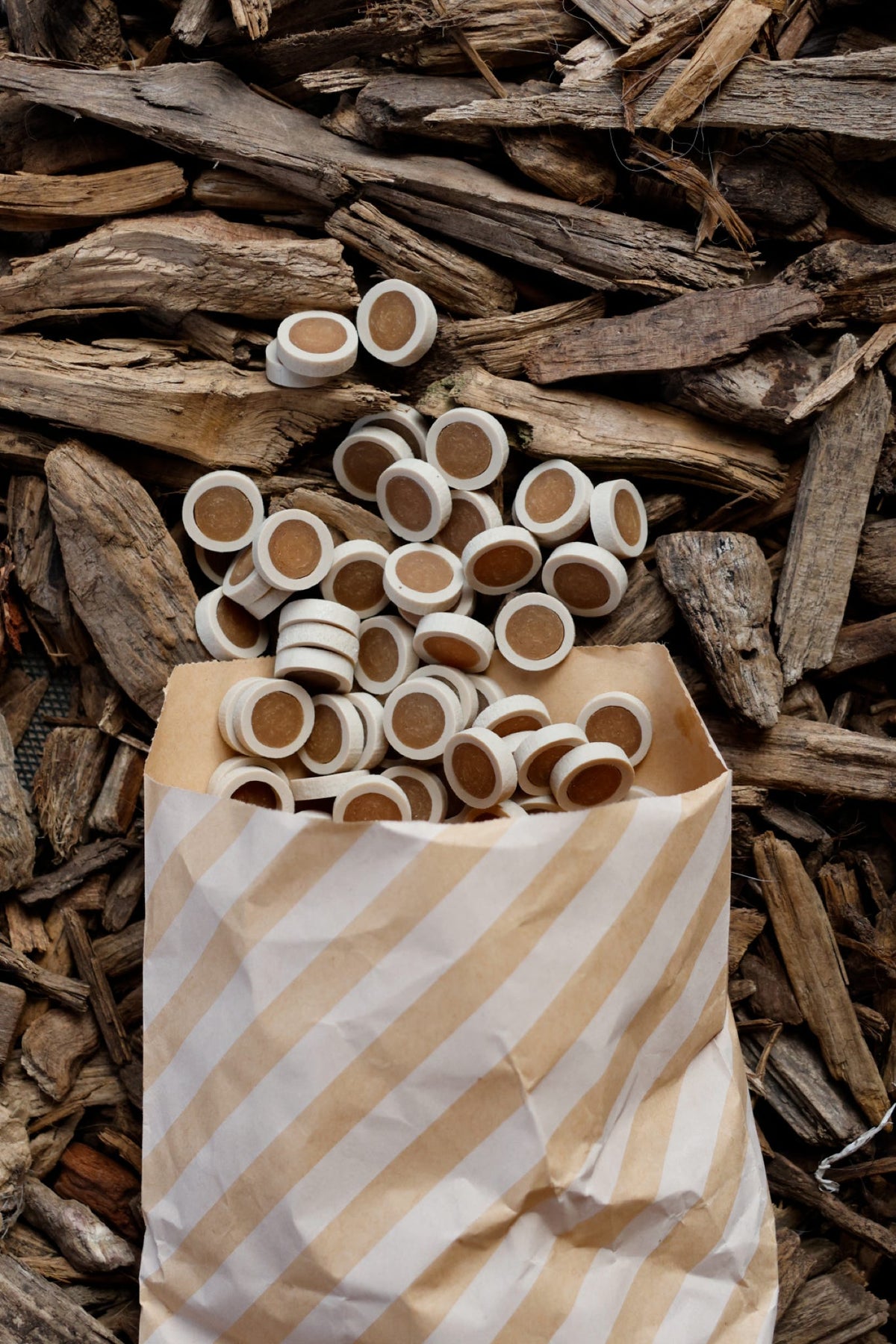 Peanut butter reward coins dog treats in a striped paper bag, displayed on a bed of natural wood chips and bark