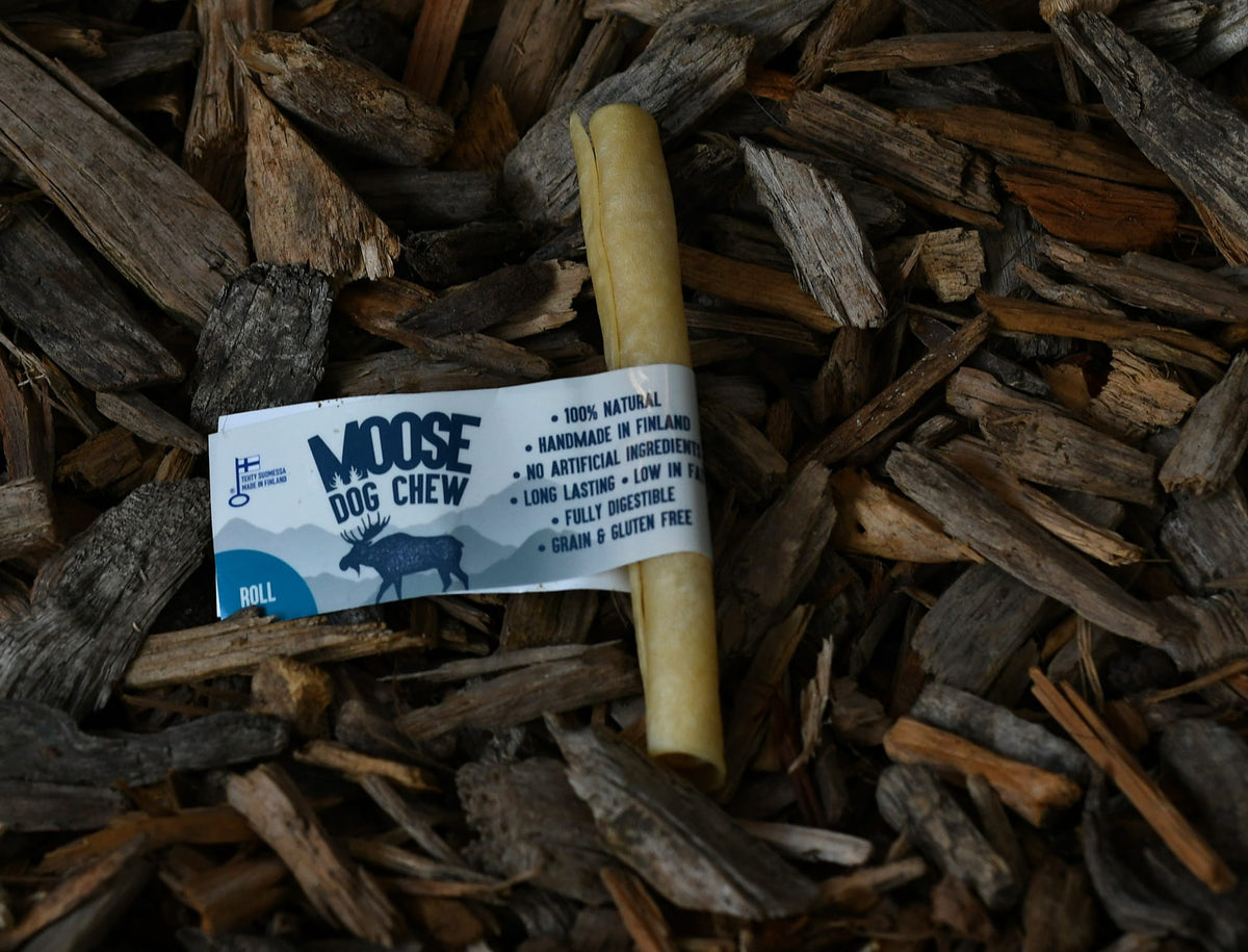 Natural moose dog chew roll with a paper label, lying on a bed of rustic wood chips.