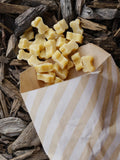 Small pale yellow bone-shaped soft cheese treats spilling from a striped paper bag onto a bed of wood chips.