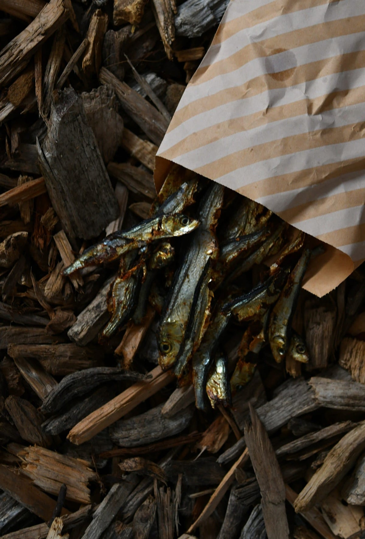 Small dried fish spilling from a brown-and-cream striped paper bag onto dark wood chips.