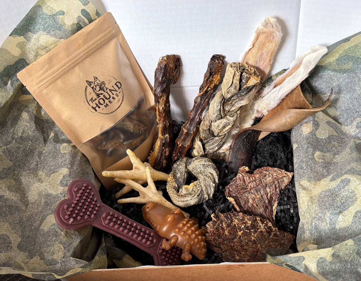 An assortment of dog treats and toys arranged in a box with camouflage tissue paper. The set includes various dried jerky-style sticks, fish skin donut, a purple textured veggie chew, rabbit ears, and a treat pouch labeled 'HOUND MESS'.