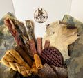 A box of dog treats in a white box with the logo 'The Hound Mess'. The treats include various skin chews, dried meat sticks, a waffle-shaped collagen treat, and a piece of animal hide, all set against a camouflage paper lining.