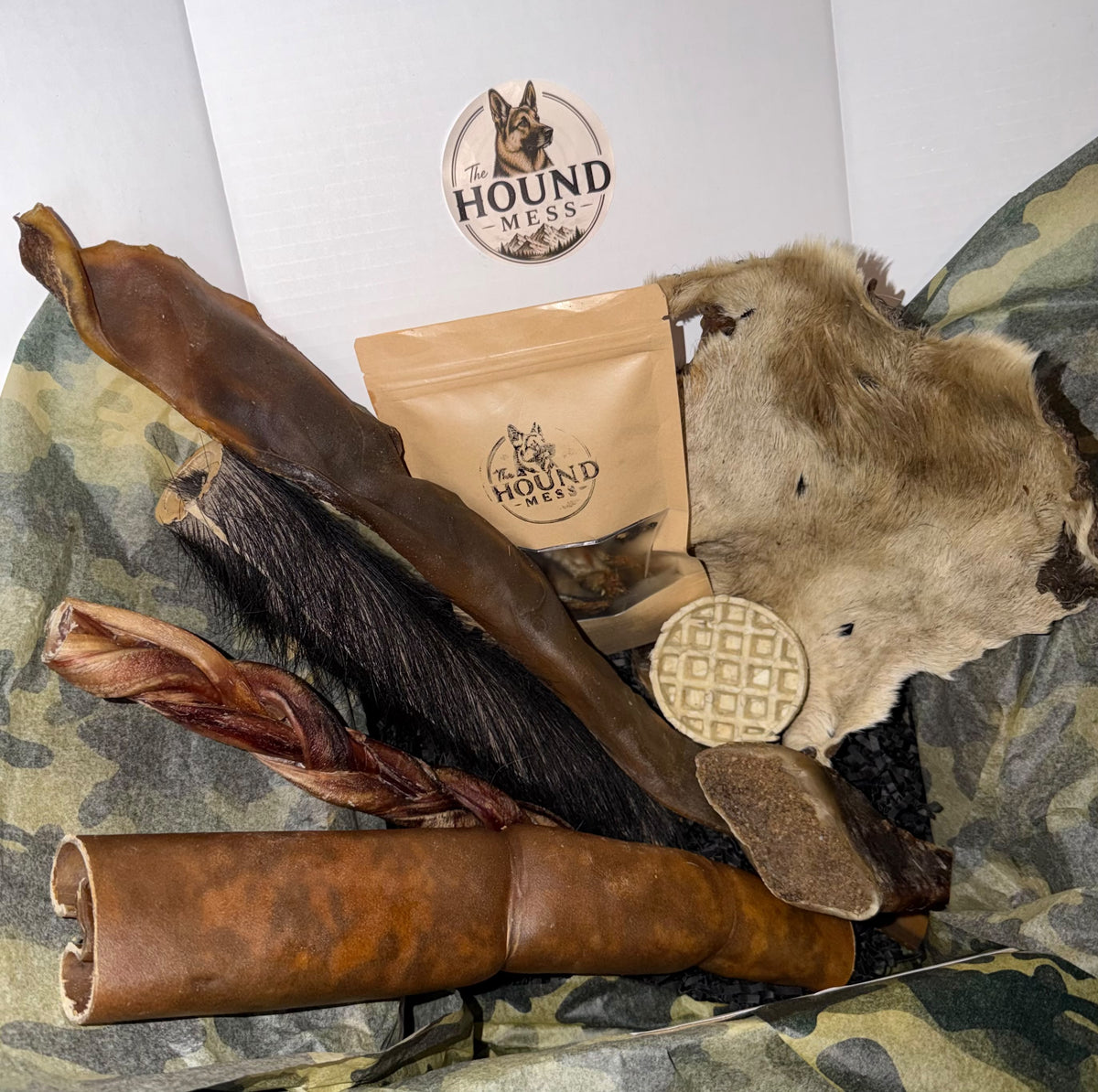 A dog chew bundle from The Hound Mess, featuring natural dog treats like a braided pizzle, a hairy skin roll and a filled hoof with the brand's logo visible.