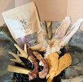 A box of dog treats and natural dog chew toys arranged in crinkle paper. Visible items include a branded treat pouch labeled 'HOUND MESS', dried jerky-style strips, puffed chicken feet, dried fish skin, dried rabbit ears, and a yak chew.