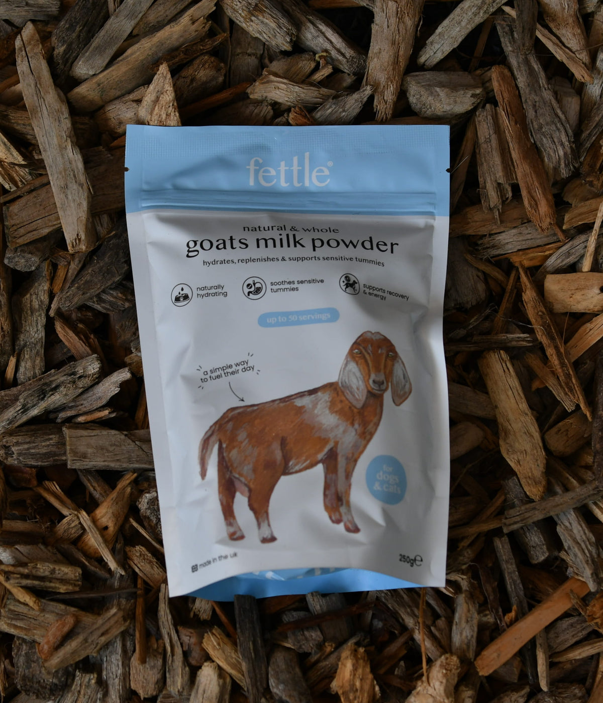 A stand-up pouch of Fettle Natural & Sourced Goats Milk Powder placed on a bed of wood chips. The pouch is white with a light blue top and bottom border, featuring a brown goat illustration and product text.