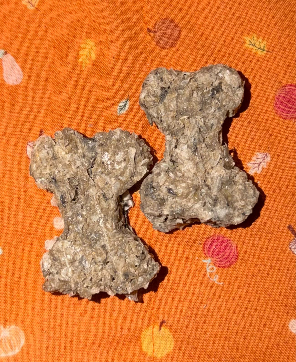 Two bone-shaped dog treats made of textured dried ingredients, placed on an orange patterned fabric background.